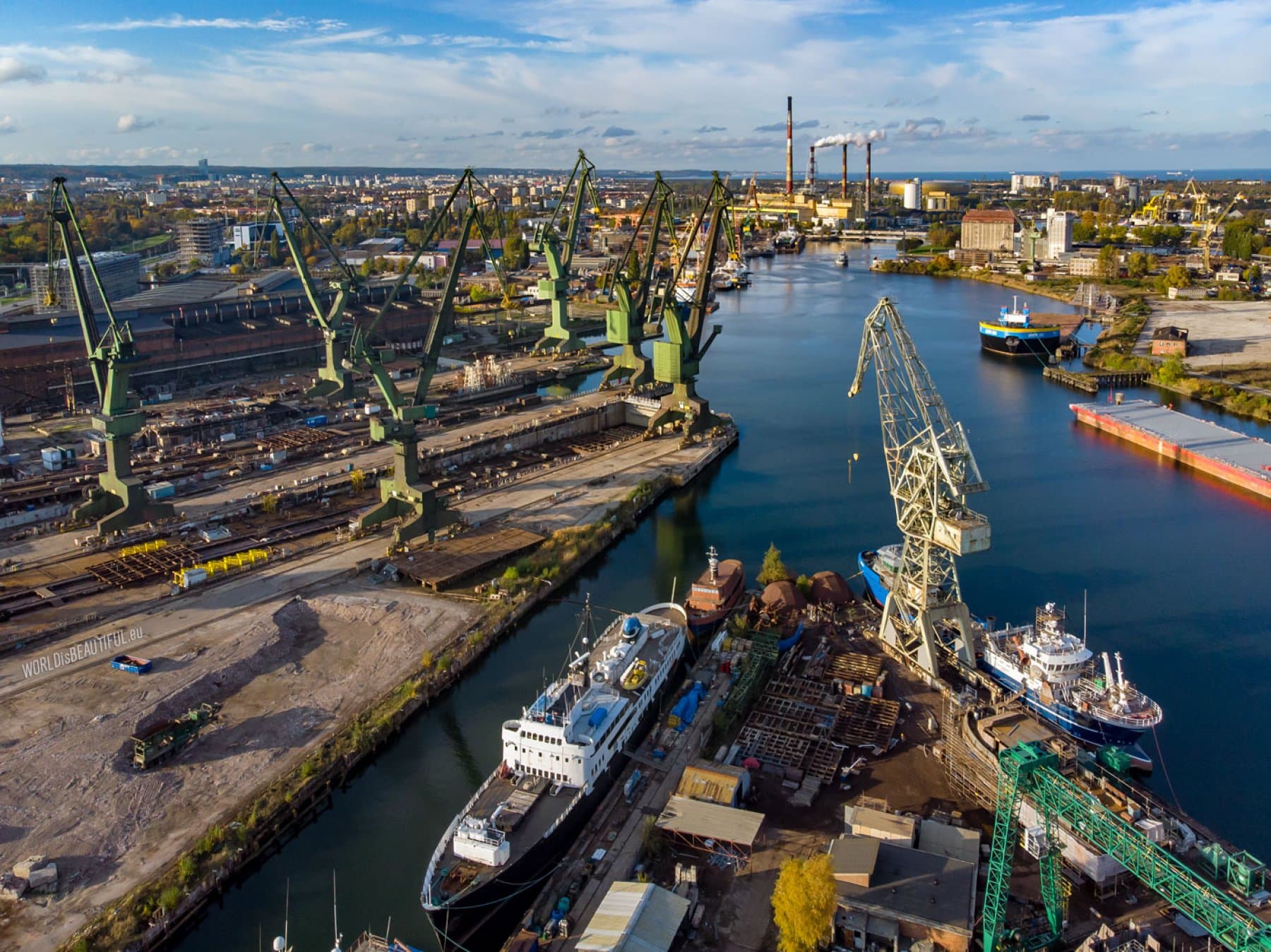 Aerial view of a bustling industrial shipyard with green cranes, docked ships, and city skyline.
