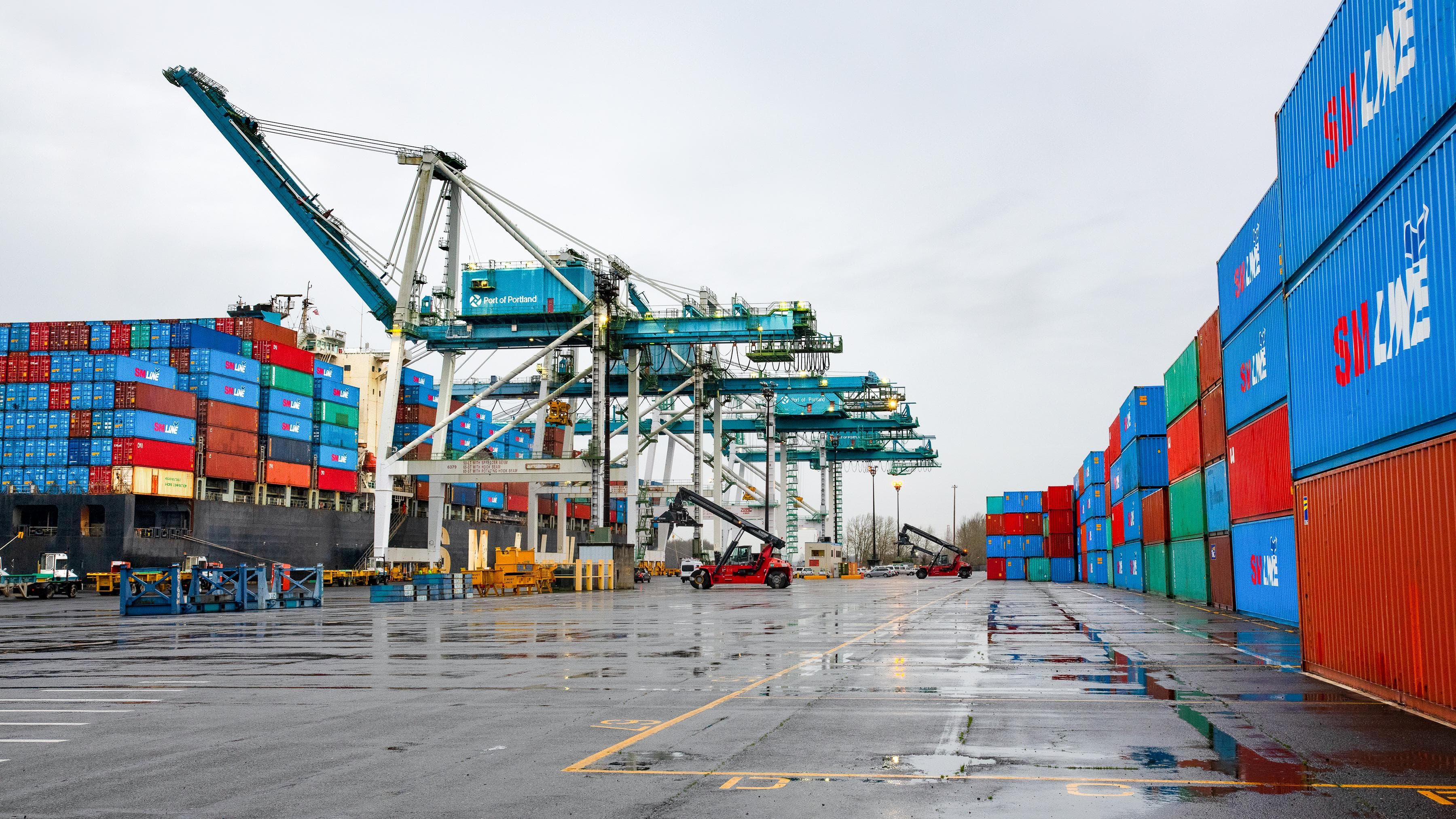 Blue gantry cranes load a container ship beside stacks of colorful containers on wet asphalt.