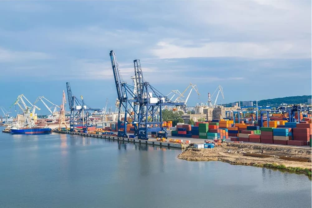 Tall blue cranes and stacks of colorful shipping containers at a busy industrial harbor.